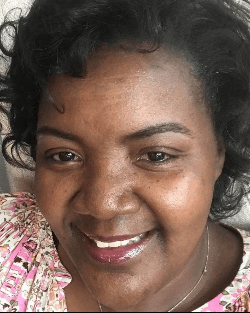 Close-up portrait of a smiling woman with short dark hair, wearing a patterned pink top and a necklace.